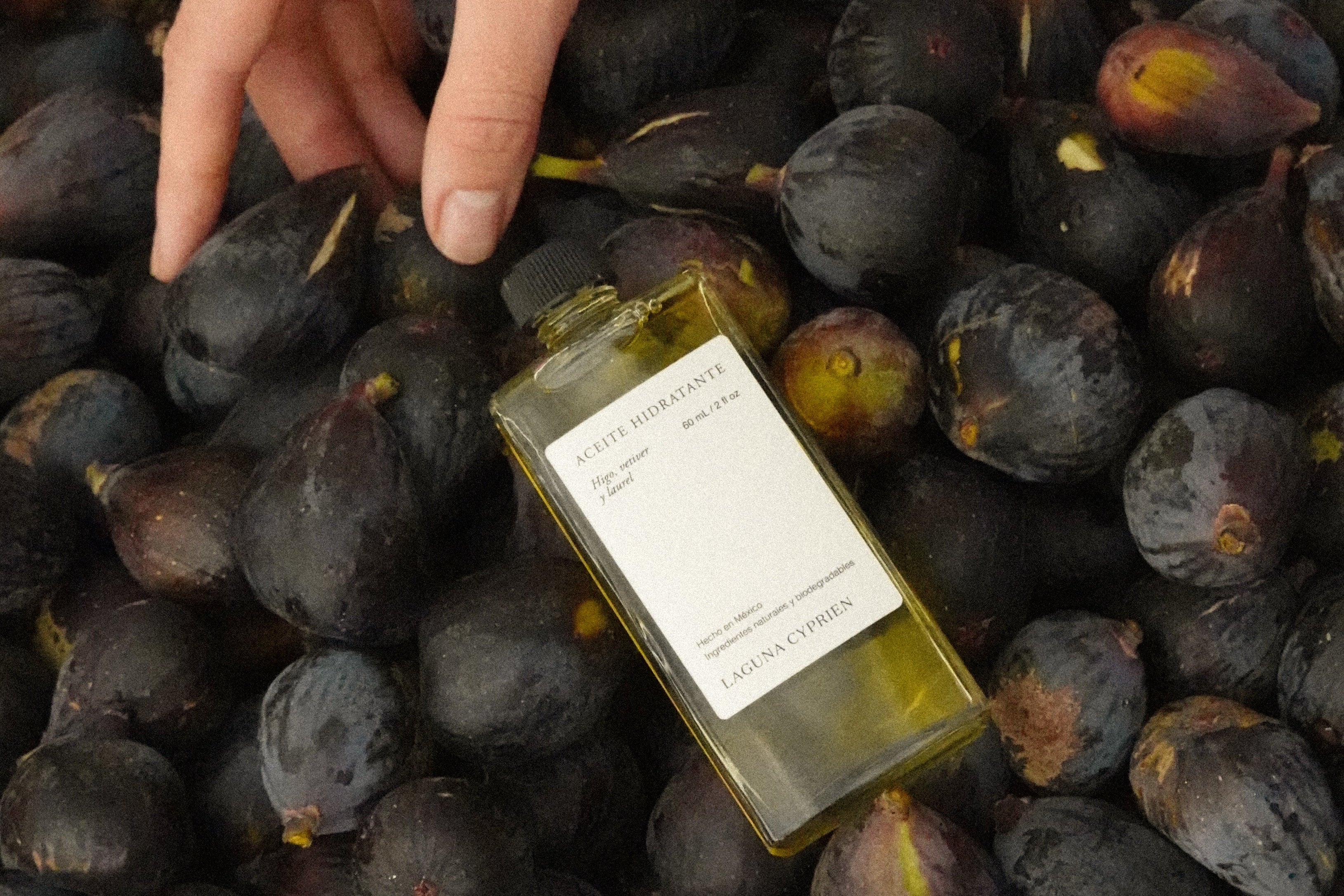 Aceite Hidratante de Laguna Cyprien acostado sobre una canasta de higos con una mano arriba.