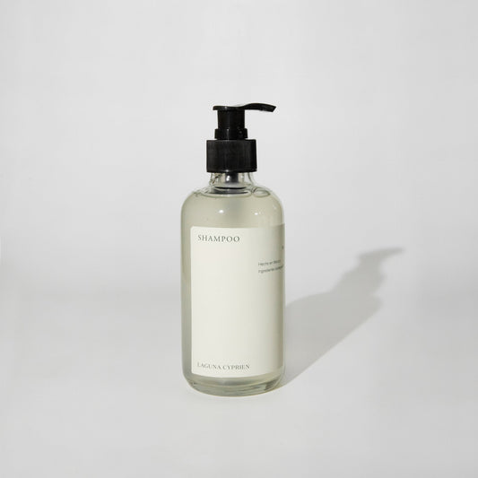Botella de cristal transparente con dosificador, etiqueta «Laguna Cyprien Shampoo Bruma Cálida», fondo blanco.